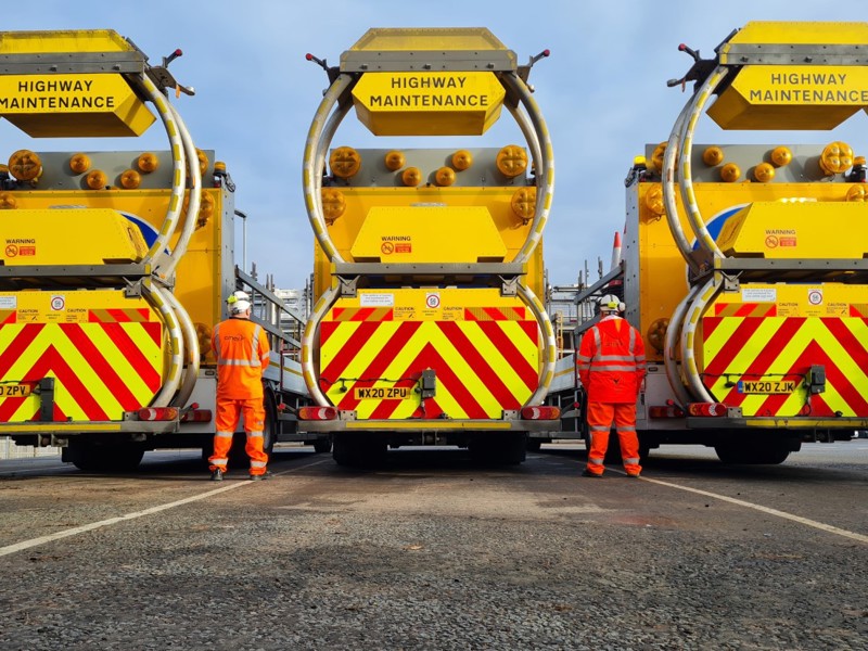 Three side by side highways maintenance vehicles, with a man in PPE stood between each one.