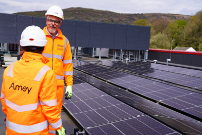 Two workers in high-visibility orange Amey PPE and hard hats stand on a rooftop solar installation, talking beside rows of solar panels with hills in the background.