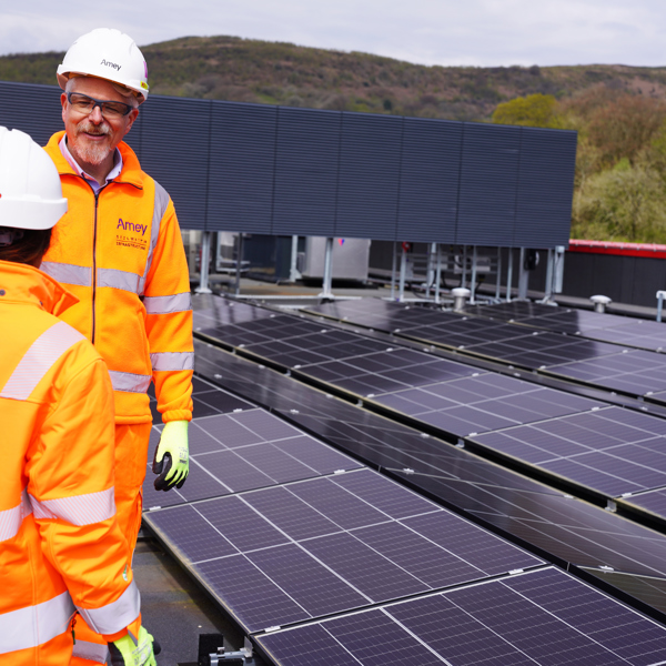 Two workers in high-visibility orange Amey PPE and hard hats stand on a rooftop solar installation, talking beside rows of solar panels with hills in the background.