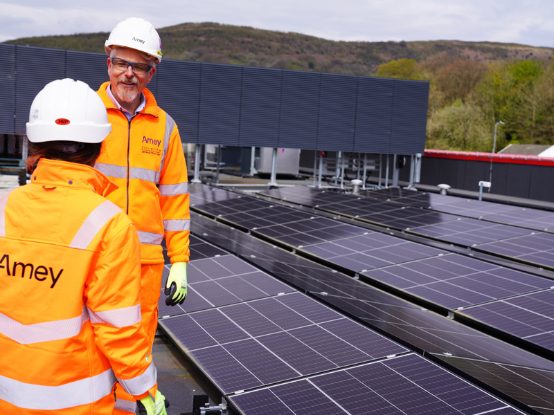 Two workers in high-visibility orange Amey PPE and hard hats stand on a rooftop solar installation, talking beside rows of solar panels with hills in the background.