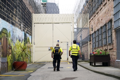 Two Amey employees in high-visibility clothing walk towards a secure gate within a prison complex, with barbed wire overhead and colourful murals and flower planters brightening the surroundings.