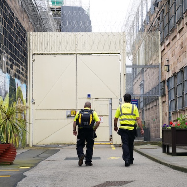 Two Amey employees in high-visibility clothing walk towards a secure gate within a prison complex, with barbed wire overhead and colourful murals and flower planters brightening the surroundings.