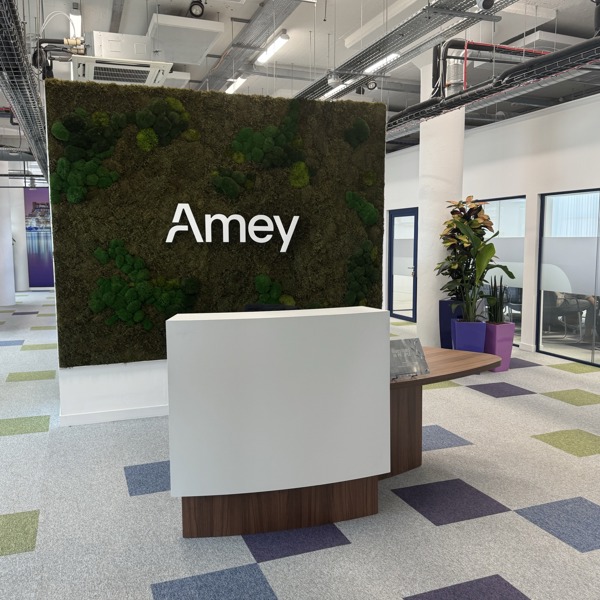 A picture of the newly refurbished Amey reception desk at Liverpool’s Matchworks, featuring a moss-covered wall with the Amey logo, a modern curved desk, and colourful square-patterned carpet tiles.