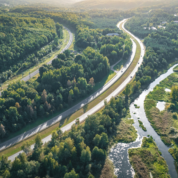 Image shows an aerial view of a highway and a railway line cutting through a lush green forest, with a winding river running alongside them