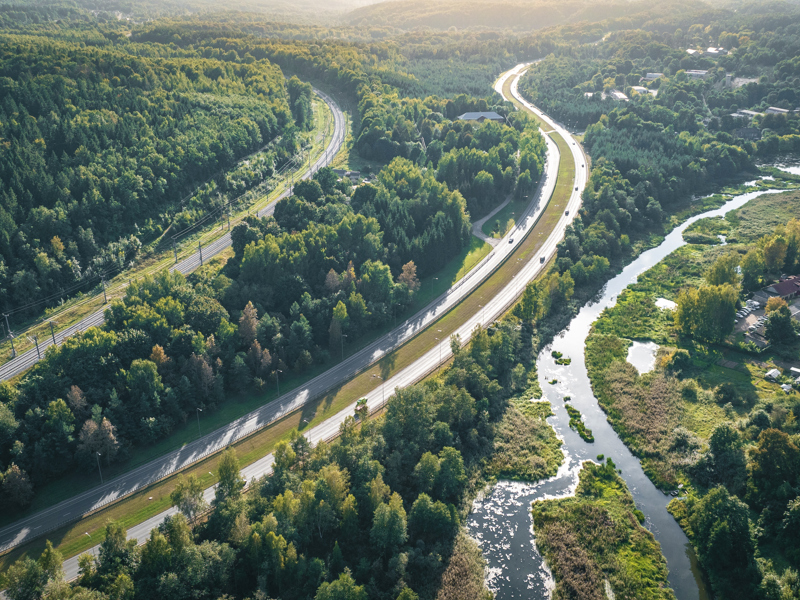 Image shows an aerial view of a highway and a railway line cutting through a lush green forest, with a winding river running alongside them
