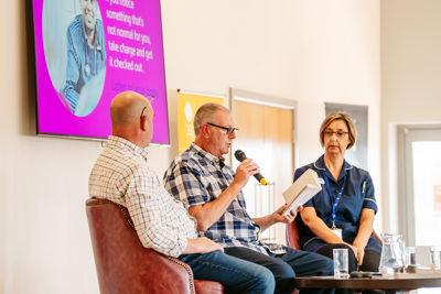 Two men and one woman sitting at a table on a stage. One man is holding a microphone and reading from a book.