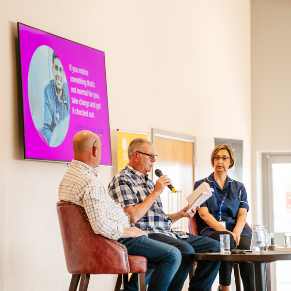 Two men and one woman sitting at a table on a stage. One man is holding a microphone and reading from a book.