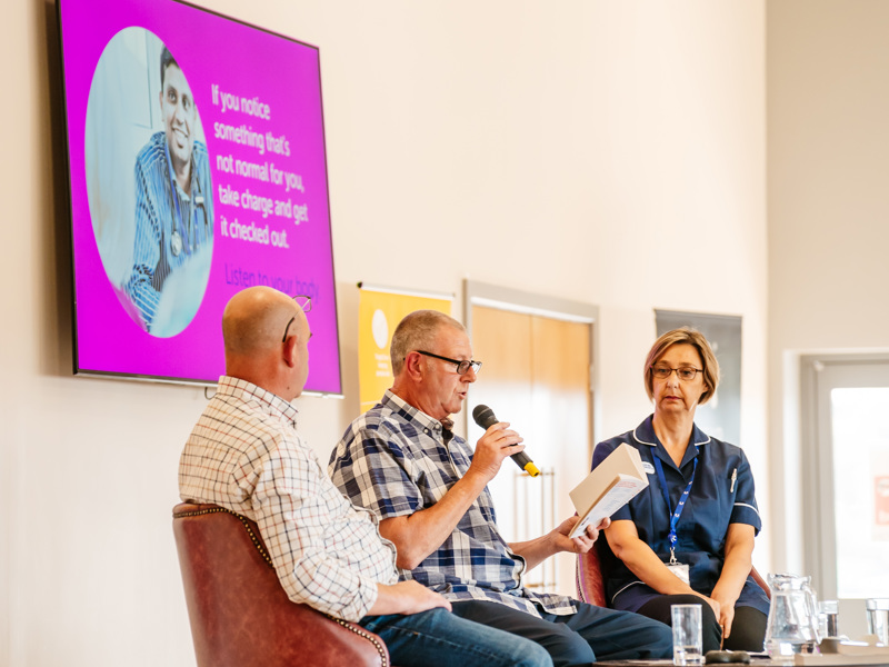 Two men and one woman sitting at a table on a stage. One man is holding a microphone and reading from a book.