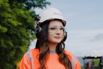 Woman wearing PPE looking into the distance. She is standing outside and a tree and body of water are visible in the background.