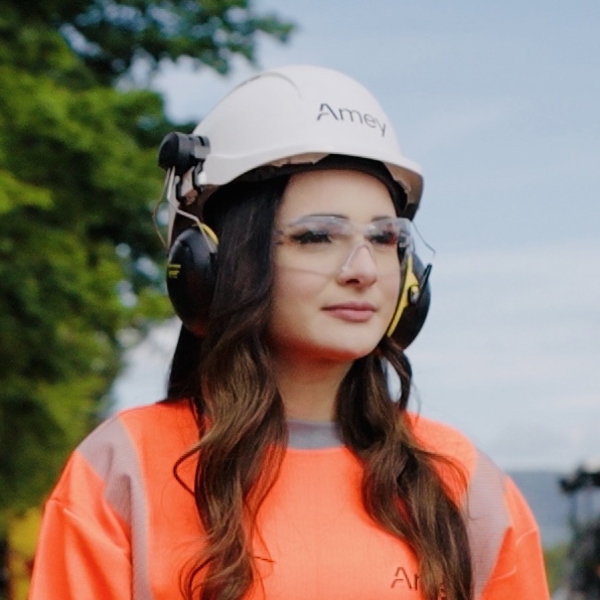 Woman wearing PPE looking into the distance. She is standing outside and a tree and body of water are visible in the background.