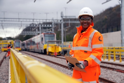 An Amey engineer wearing high-visibility orange PPE and a hard hat stands on a railway site holding a clipboard, with trains and overhead lines visible in the background.