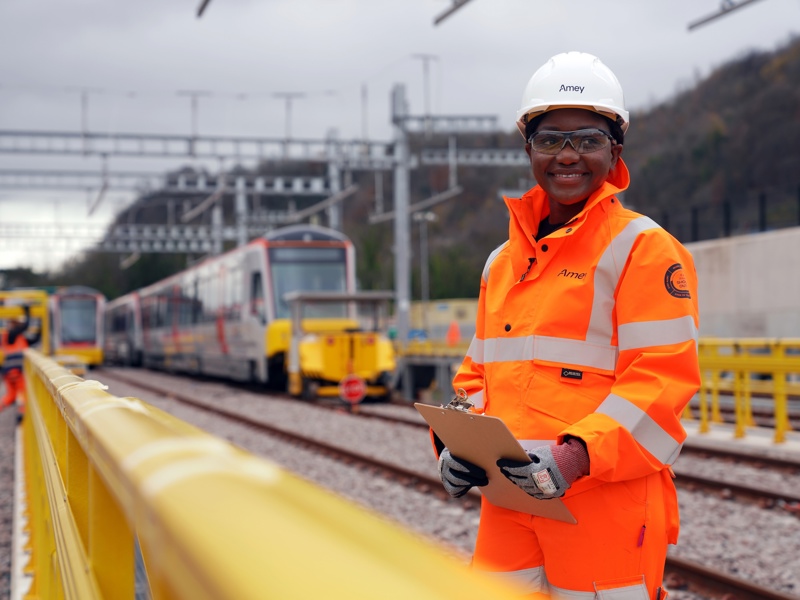 An Amey engineer wearing high-visibility orange PPE and a hard hat stands on a railway site holding a clipboard, with trains and overhead lines visible in the background.