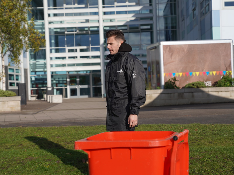Image of a male Amey employee, stood on grass, behind an orange bin. 