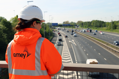 Image of a female Amey employee wearing PPE, standing with their back o the camera on a bridge.