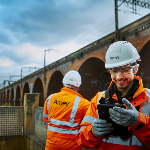 Two Amey rail workers in high-vis gear inspect infrastructure near a brick viaduct, with one smiling while using a tablet.